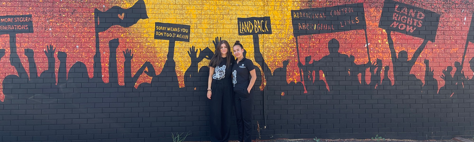 Project officers Tasha Cole and Taleah Carson in front of an Indigenous mural. 