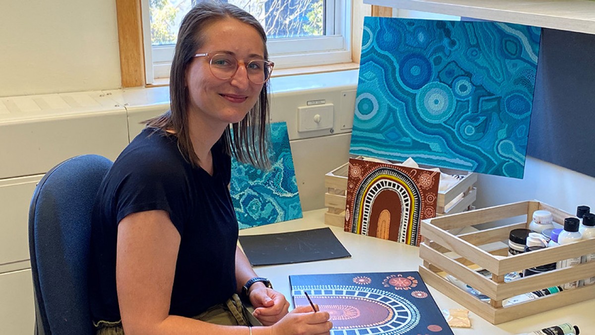 Roxy Jones is sitting at a desk painting Aboriginal artwork, with other canvases around her. 