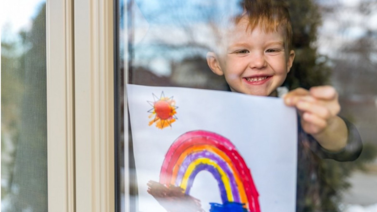 A child holds a picture of a rainbow behind a window.